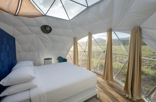 Cozy interior of a geodesic dome with a bed, large windows, and scenic views of nature outside.